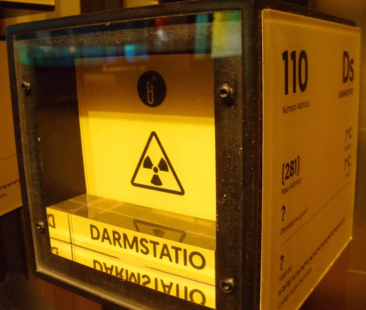 A glass display cube in a museum, lit yellow, holding only a yellow card with a radiation warning sign and a block engraved “Darmstatio”; the side panel reads 110, Ds, (281)