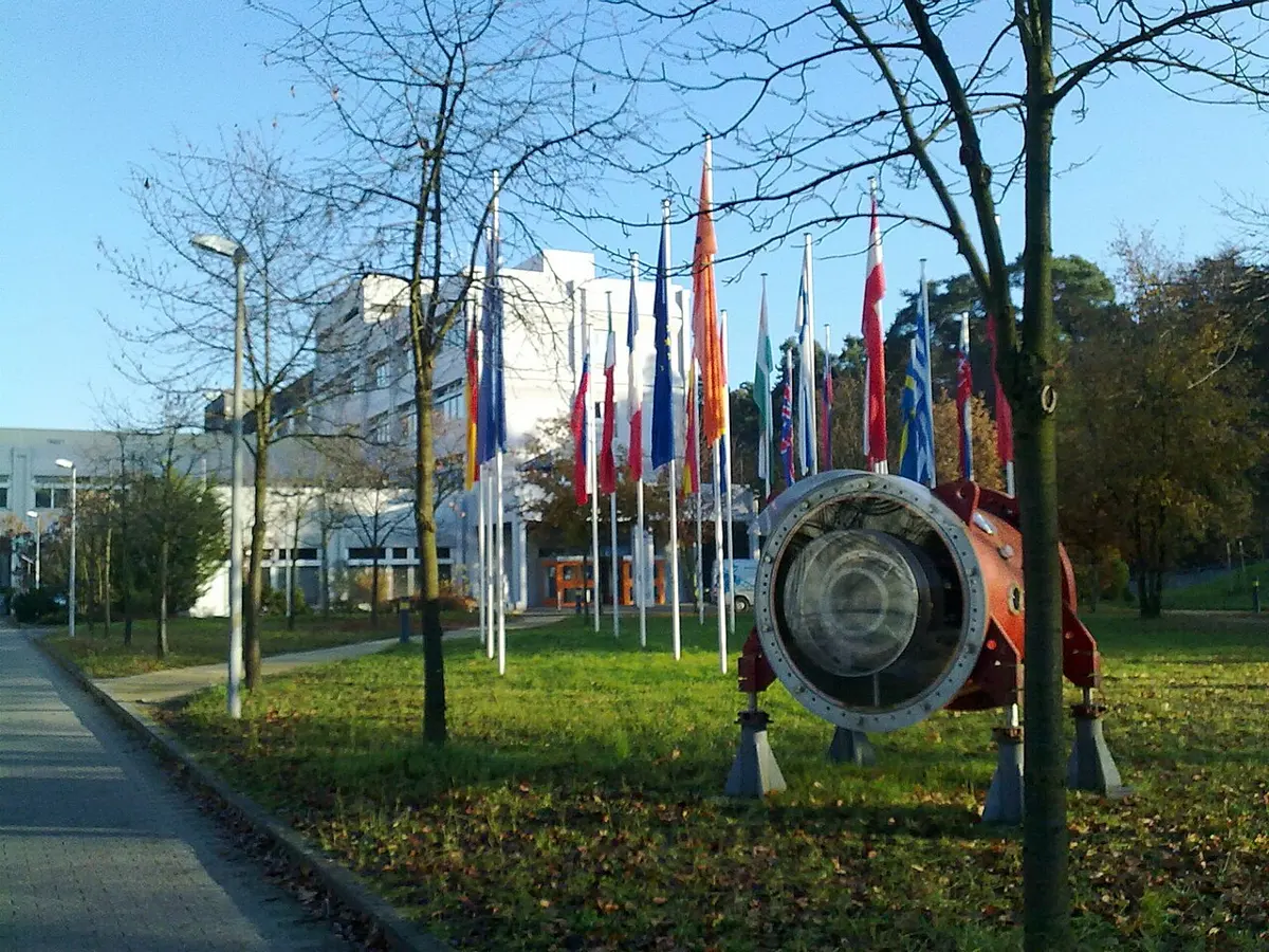A lawn with a row of flags of many countries in front of a long white office building; in the front, a large round red-and-silver machine part stands on metal legs