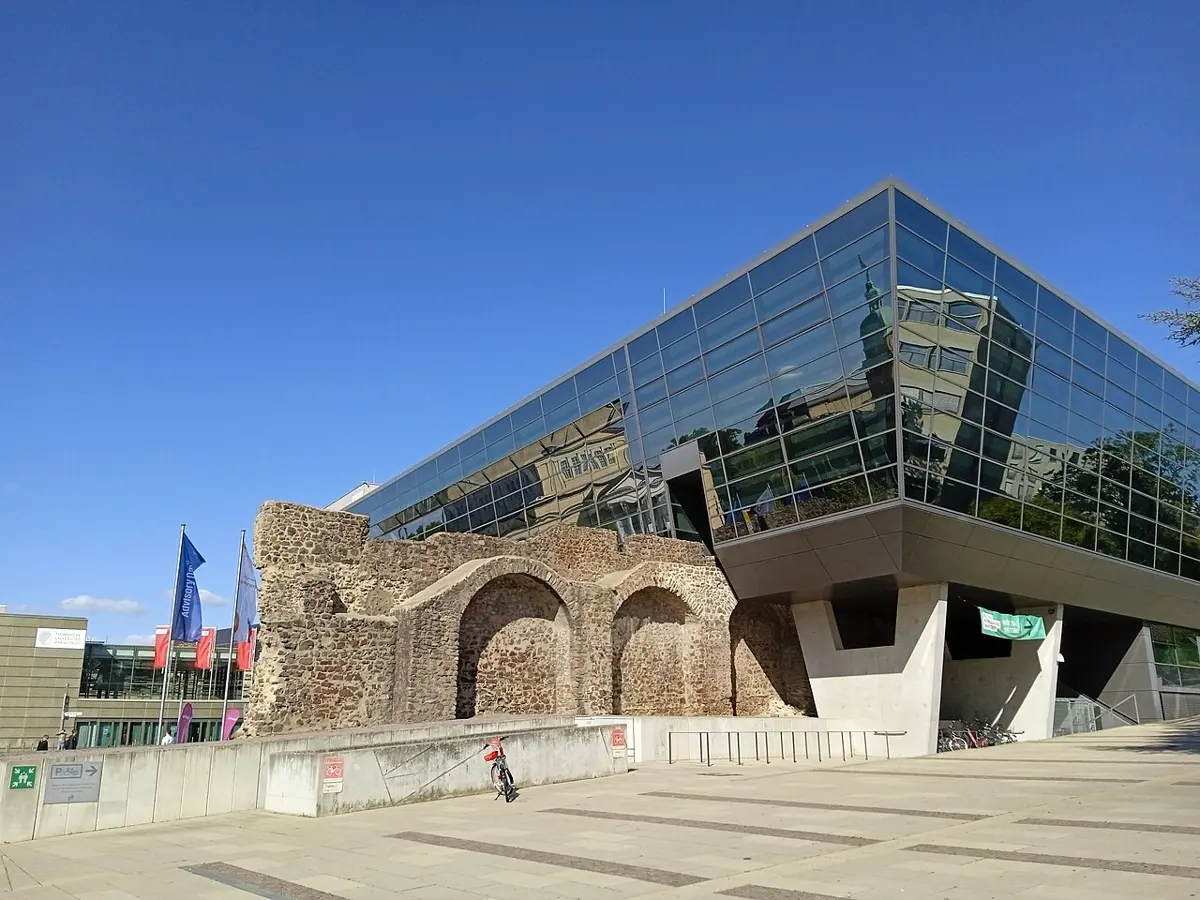 A modern building with a sloping glass front and a gray stone side, standing next to the remains of an old stone wall with arches, under a blue sky