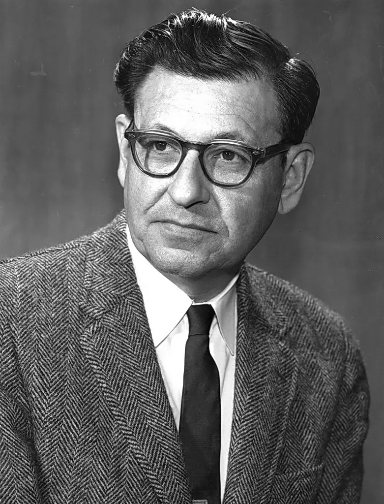 A black-and-white portrait of a man with dark wavy hair and thick-rimmed glasses, in a jacket and tie