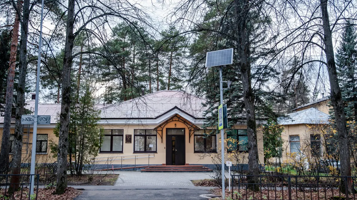 A single-storey yellow building with a peaked porch and a dark door, behind a low iron fence among tall pine and birch trees