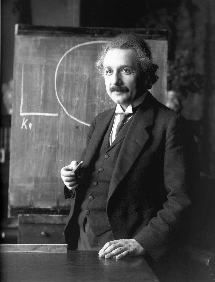 A black-and-white photo of a man with wavy hair and a mustache, in a three-piece suit, holding a piece of chalk in front of a blackboard