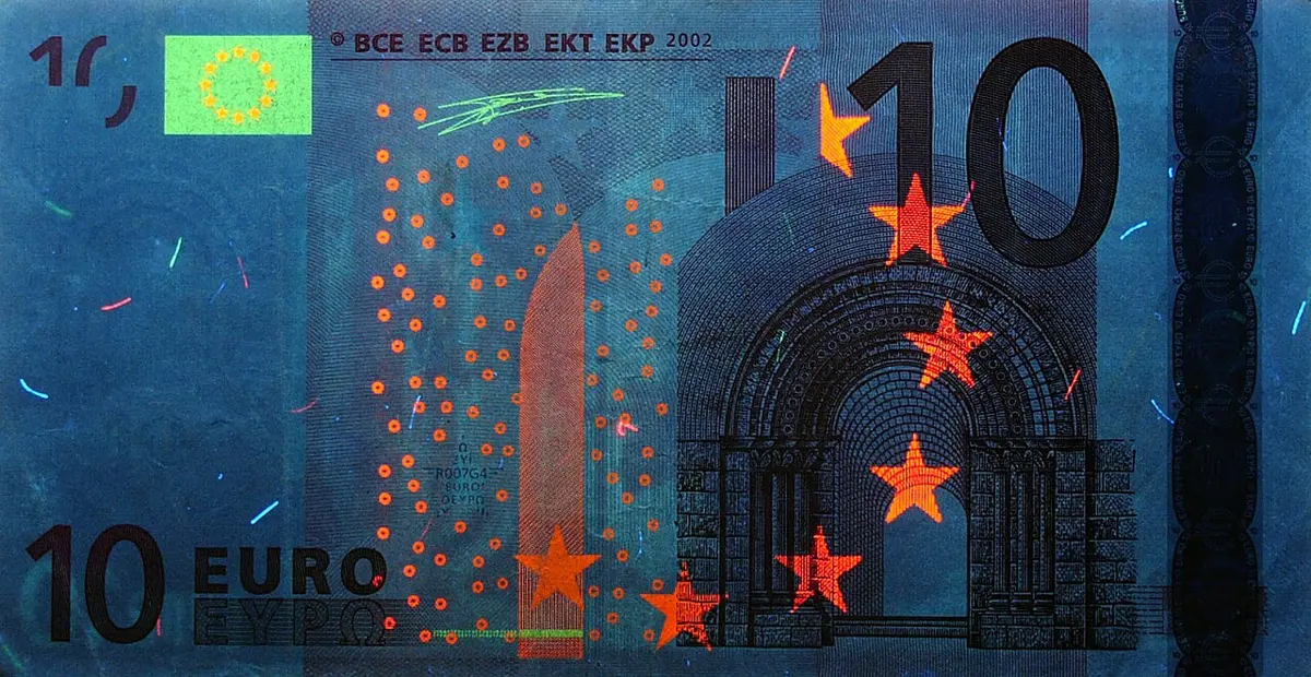 A 10 euro banknote under ultraviolet light: the note looks dark blue, with orange-red stars, a band of orange-red dots and short colored fibers glowing