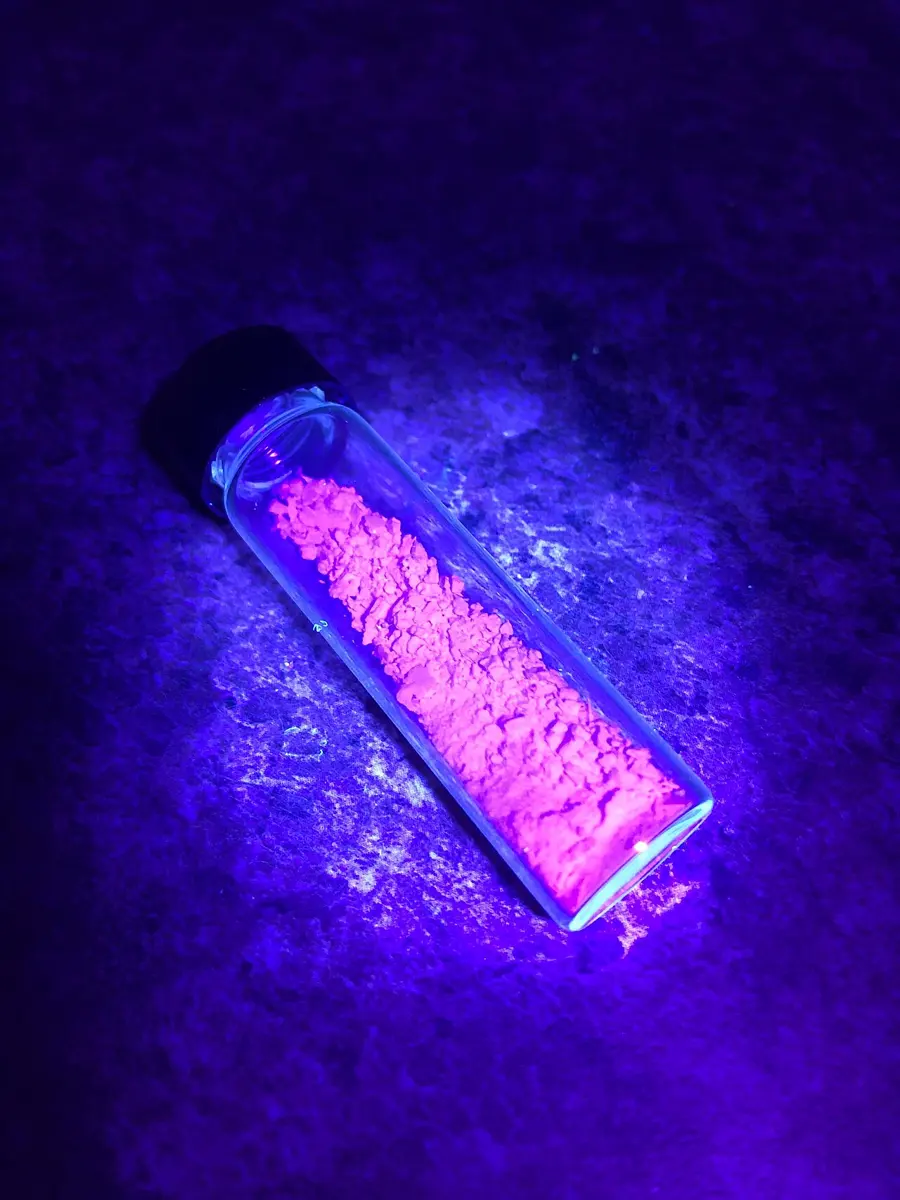 A small glass vial of powder glowing bright pink-red in the dark, lit by a violet ultraviolet lamp