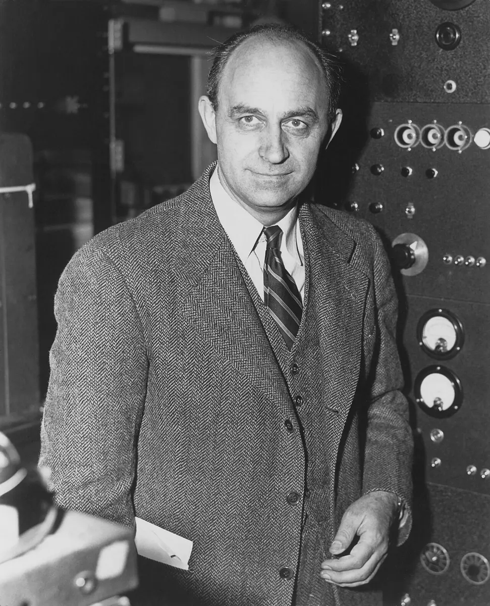 A black-and-white photo of a man with receding hair, in a tweed jacket and striped tie, standing in front of a panel of dials and switches