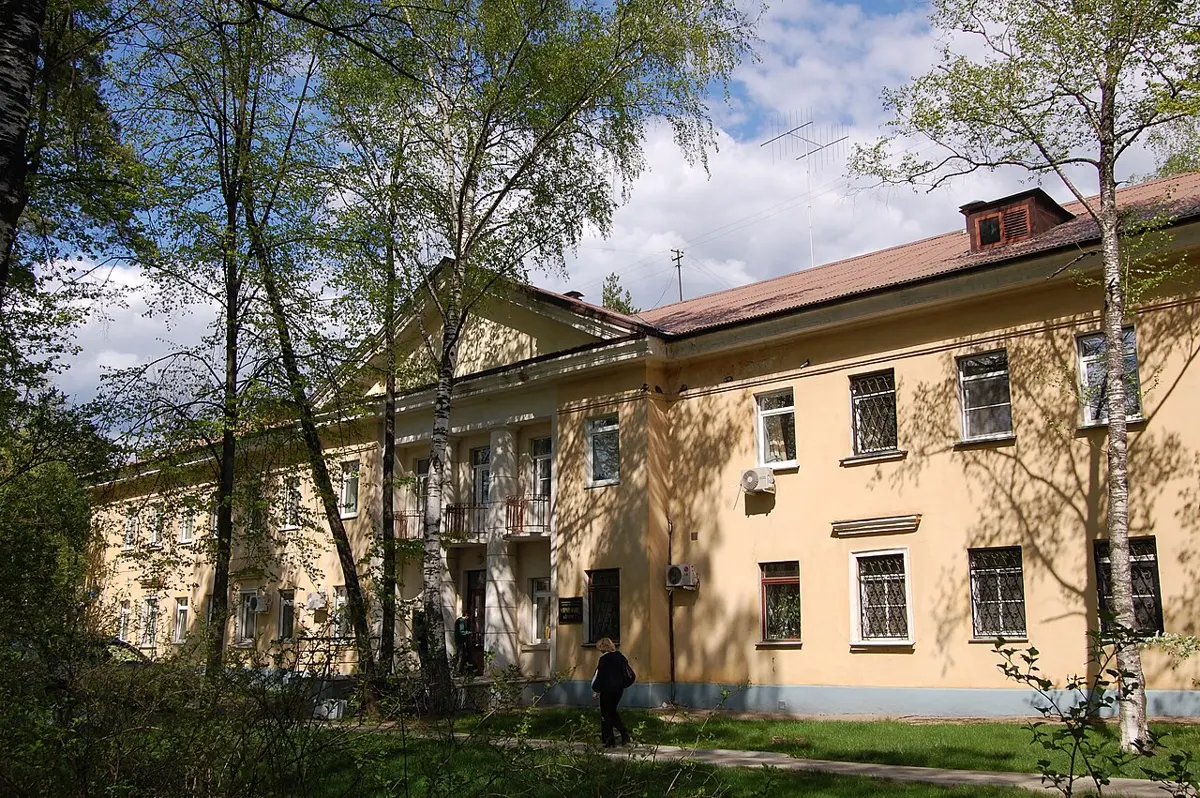 A long, pale yellow two-storey building with white window frames behind trees and a lawn, under a blue sky
