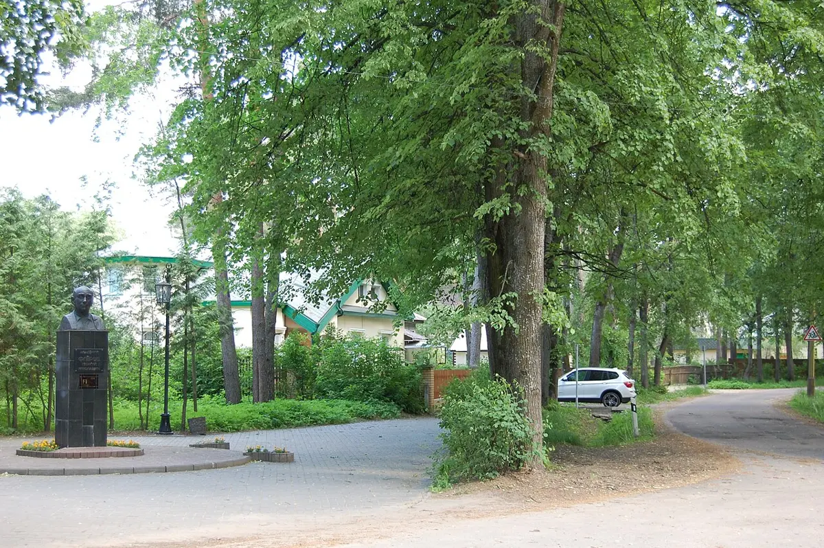 A quiet tree-lined road with a small paved square on the left, where a bust stands on a dark pedestal; houses with green roofs behind the trees