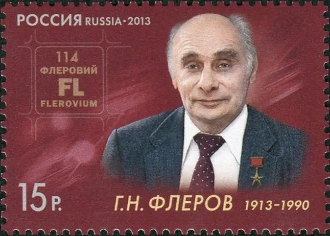 A red Russian postage stamp from 2013 with a portrait of an elderly, balding man in a suit and tie, and a periodic-table box reading 114, Fl, flerovium