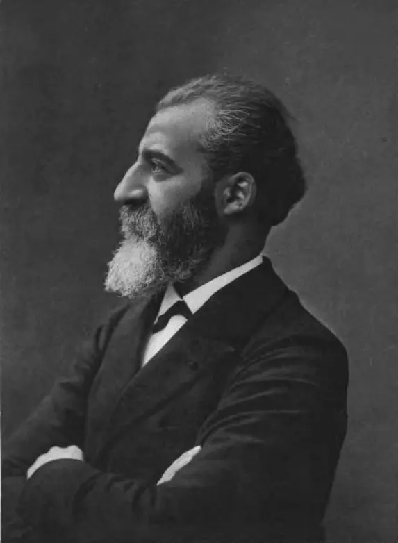 A black and white side view of a man with short dark hair and a gray-white beard, in a dark suit and bow tie, his arms folded