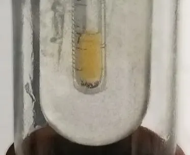 A small glass tube standing inside a larger, frosted glass vessel, with a little yellow liquid at its bottom
