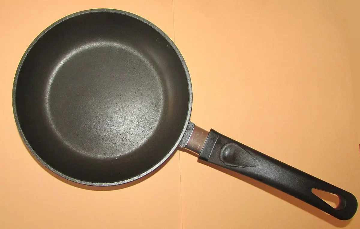 A frying pan seen from above on an orange background, its inside coated dark gray, with a black plastic handle