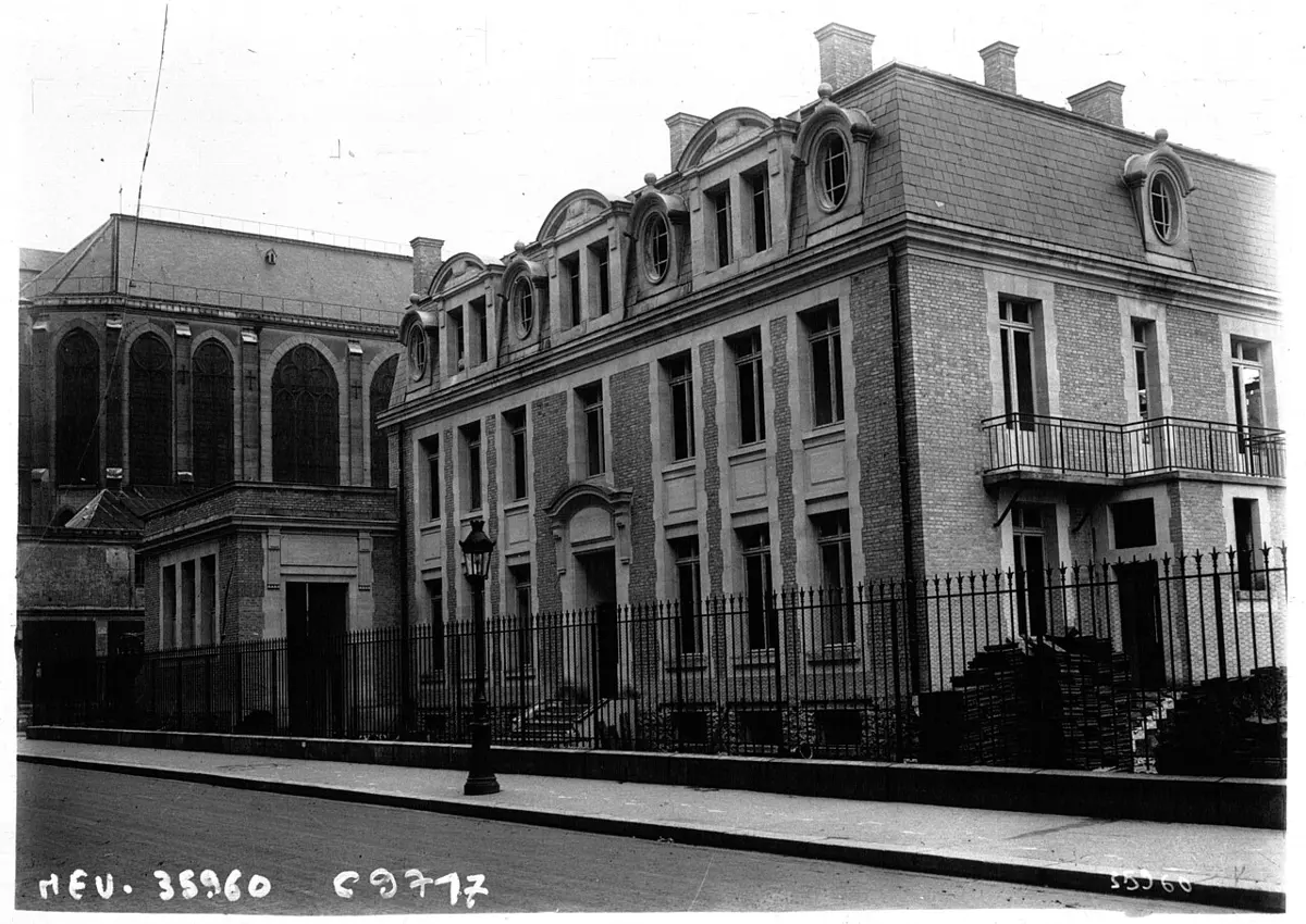 An old black and white photograph of a three-storey brick and stone building with round windows in its slate roof, behind a tall iron fence on a quiet street, with stacks of bricks by the wall