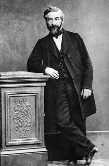 An old black-and-white photograph of a bearded man in a dark suit and waistcoat, standing with one elbow on a carved pedestal