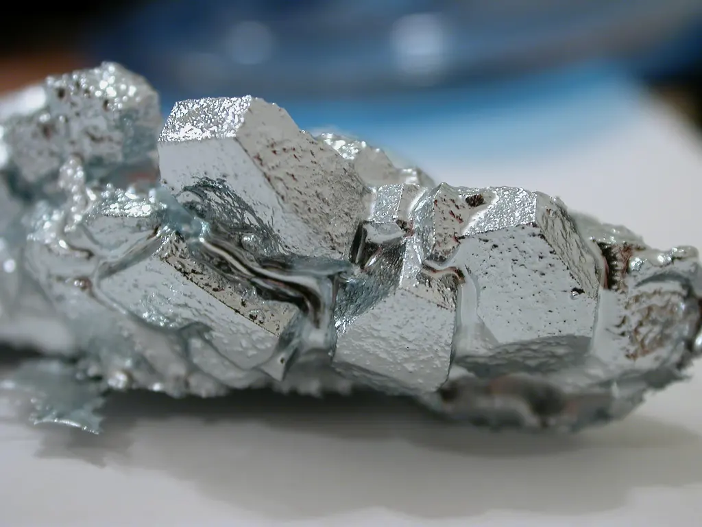 A lump of shiny, silvery metal made of blocky crystals with flat faces and sharp edges, on a white surface