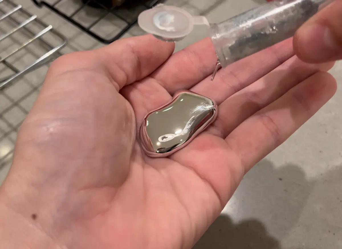 A shiny, mirror-like puddle of liquid metal in the palm of a hand, poured from a small plastic tube