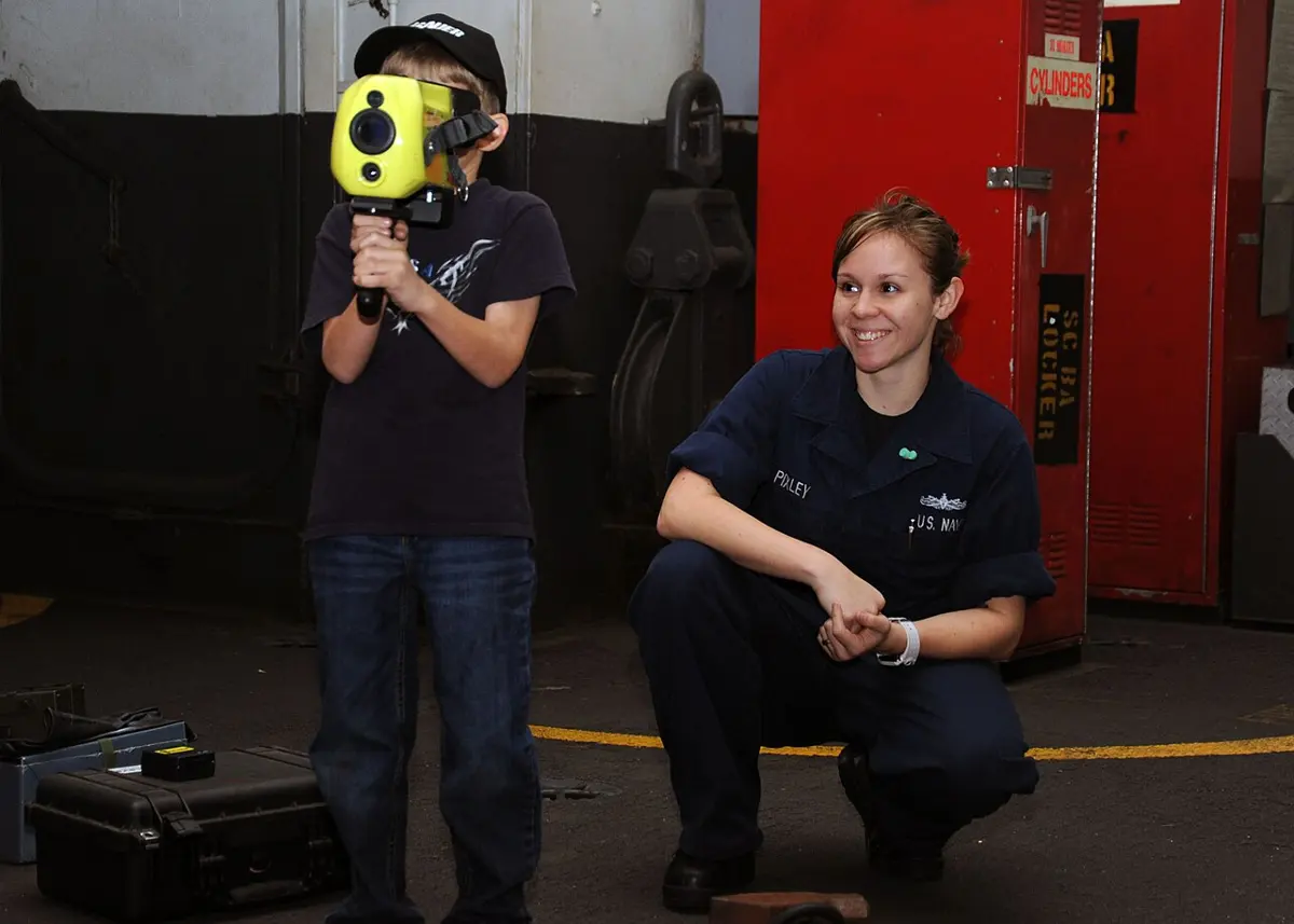 A boy holds a yellow thermal camera up to his eyes, its round, dark lens pointing towards us, while a smiling woman in a navy uniform kneels beside him