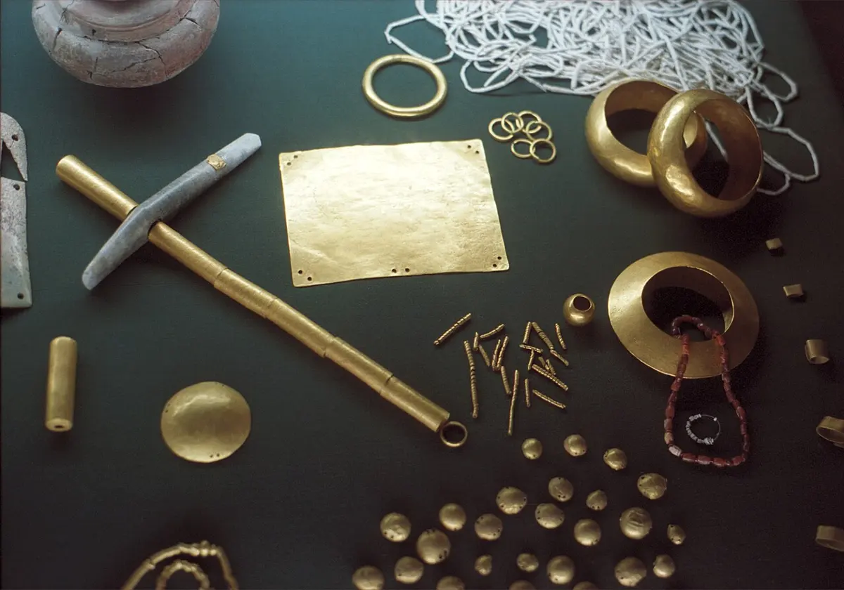 A museum case of ancient gold objects on dark green cloth: bracelets, rings, small round plaques, a rectangular plate, a long gold-covered staff and many little beads