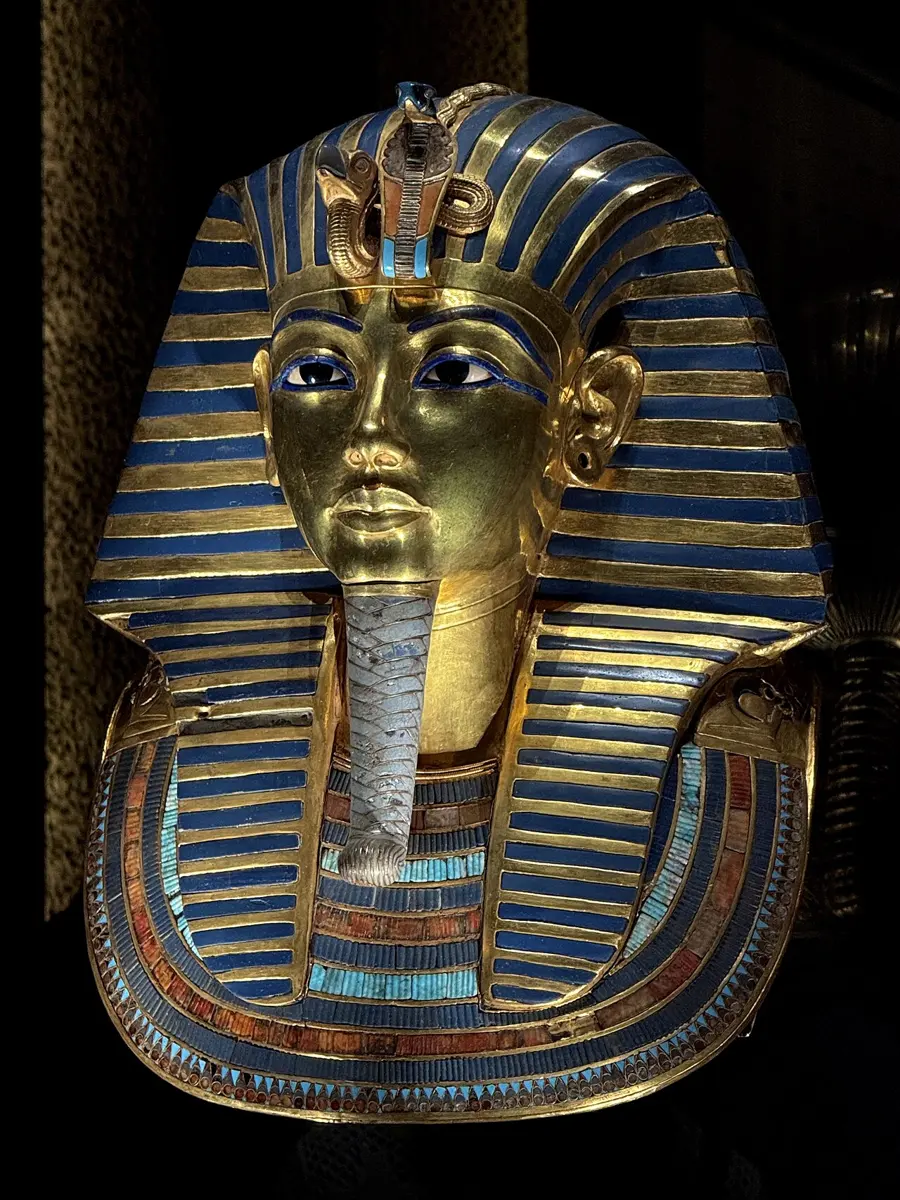 The golden funeral mask of an Egyptian king, with a striped headdress of gold and blue glass, a cobra and vulture on the brow and a long braided beard