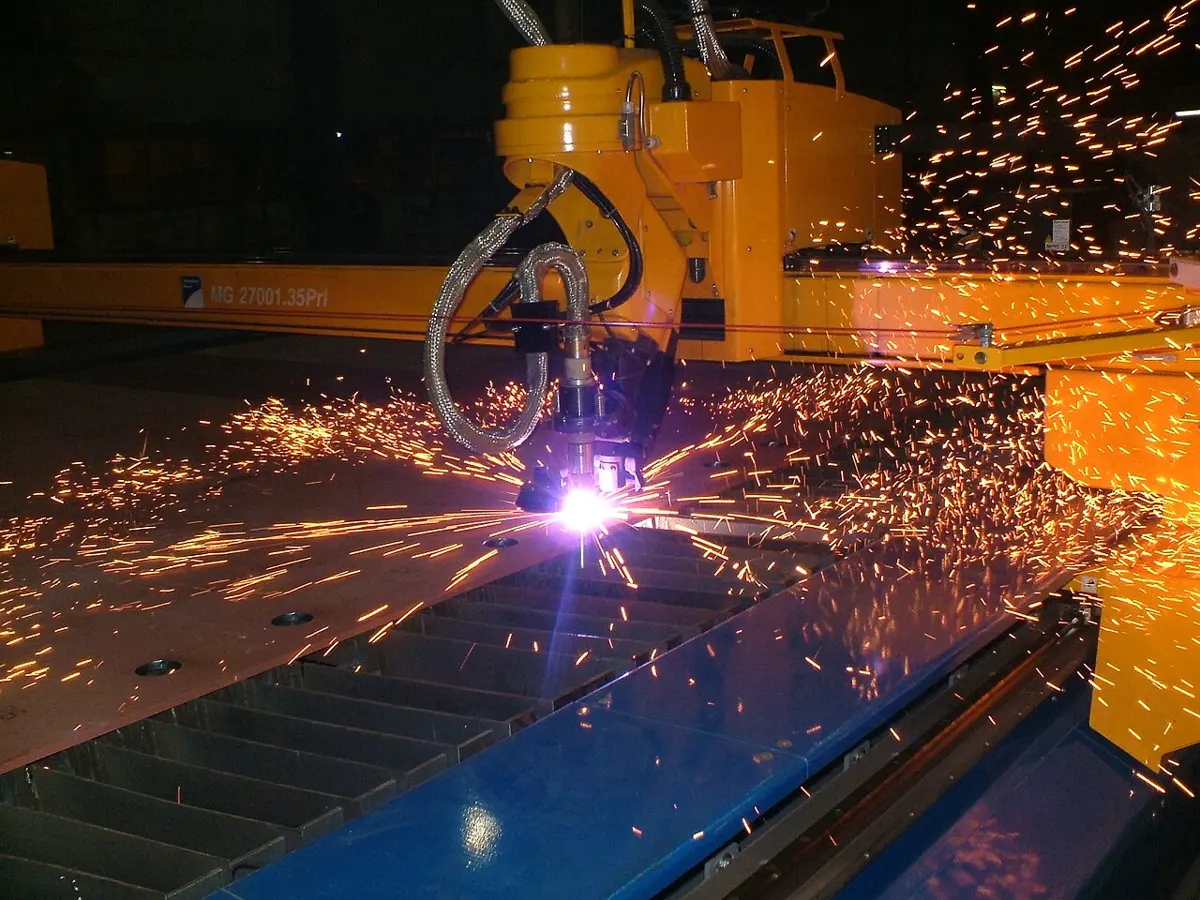 A yellow cutting machine moving over a large steel plate in a dark factory; a bright violet-white spark at the torch’s tip sends showers of orange sparks across the plate