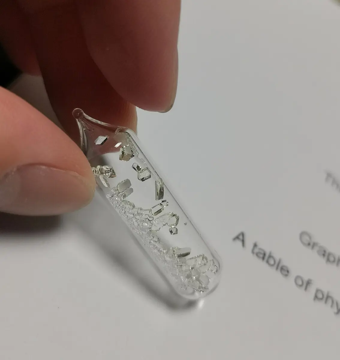 Fingers holding a small sealed glass tube containing clear, colorless crystals, above a printed page