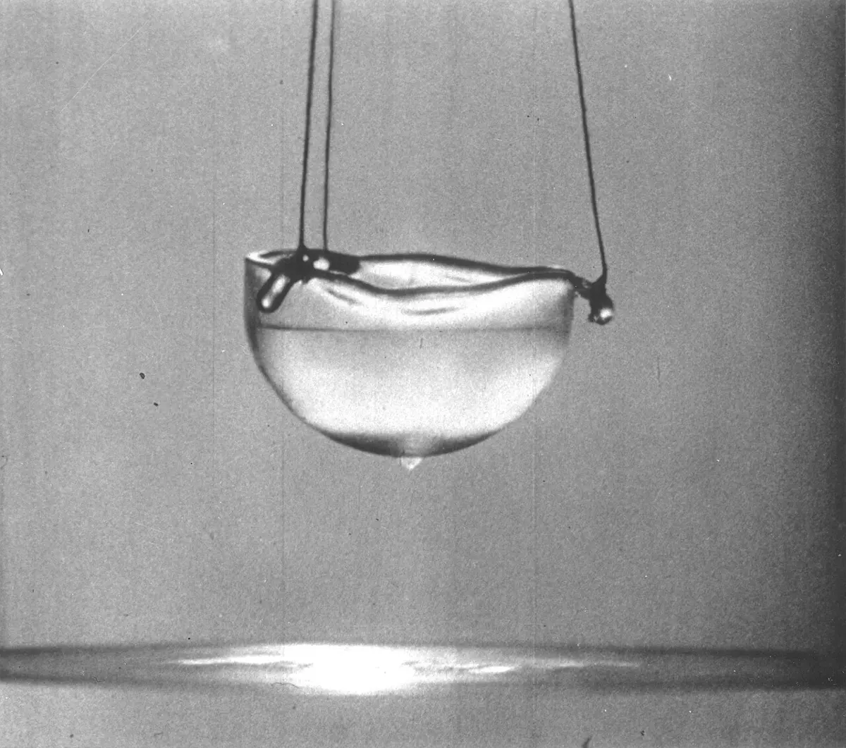 Black-and-white photo of a small glass cup of clear liquid hanging on threads, with a drop forming underneath its rounded bottom