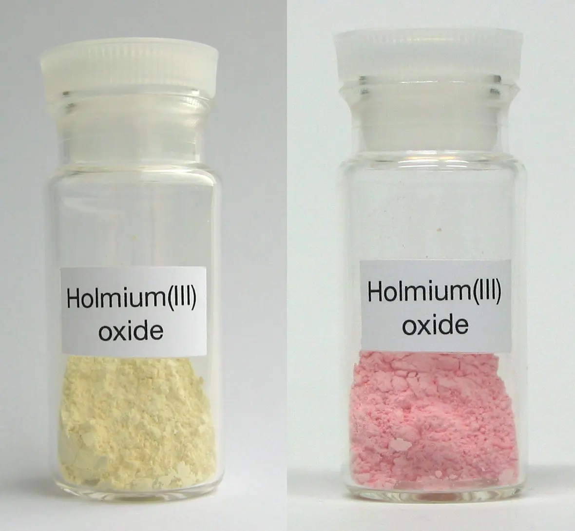 Two identical small glass bottles labeled Holmium(III) oxide; the powder in the left one is pale yellow, in the right one pink