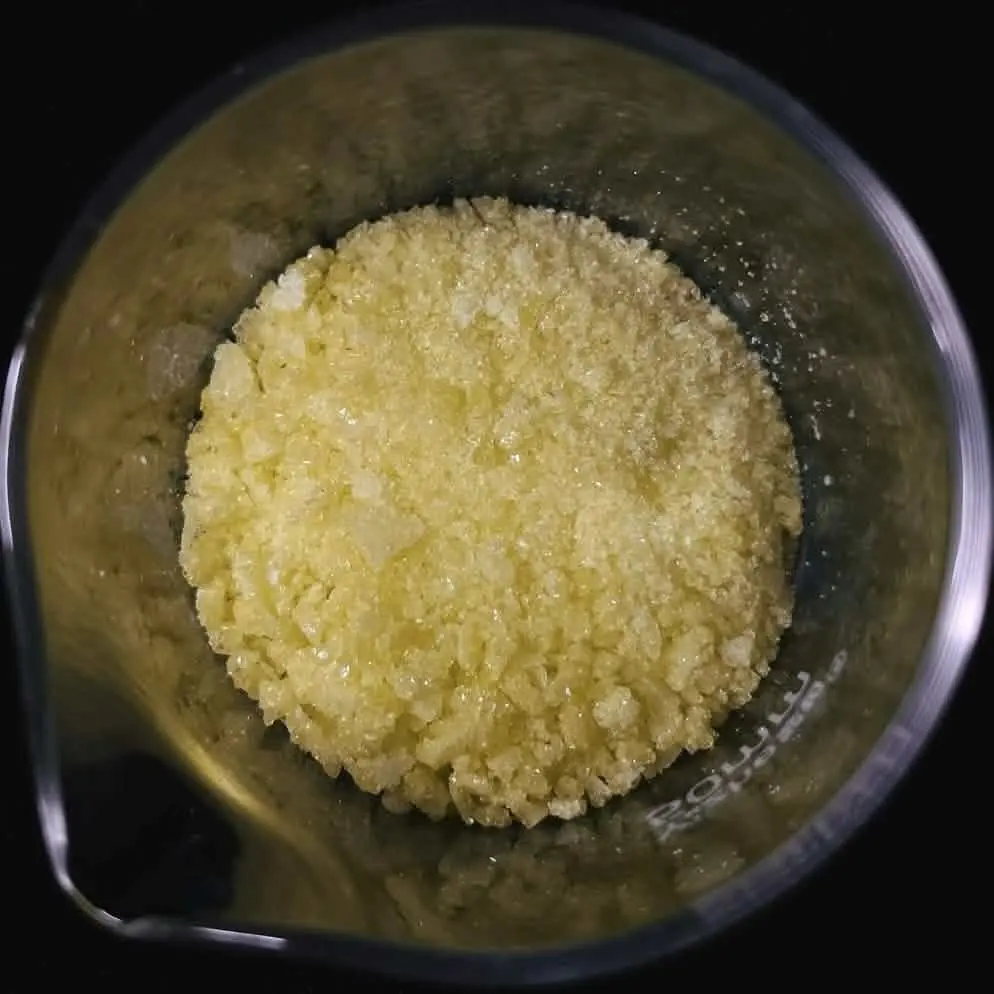 Pale yellow crystals filling the bottom of a glass beaker, seen from above against a dark background