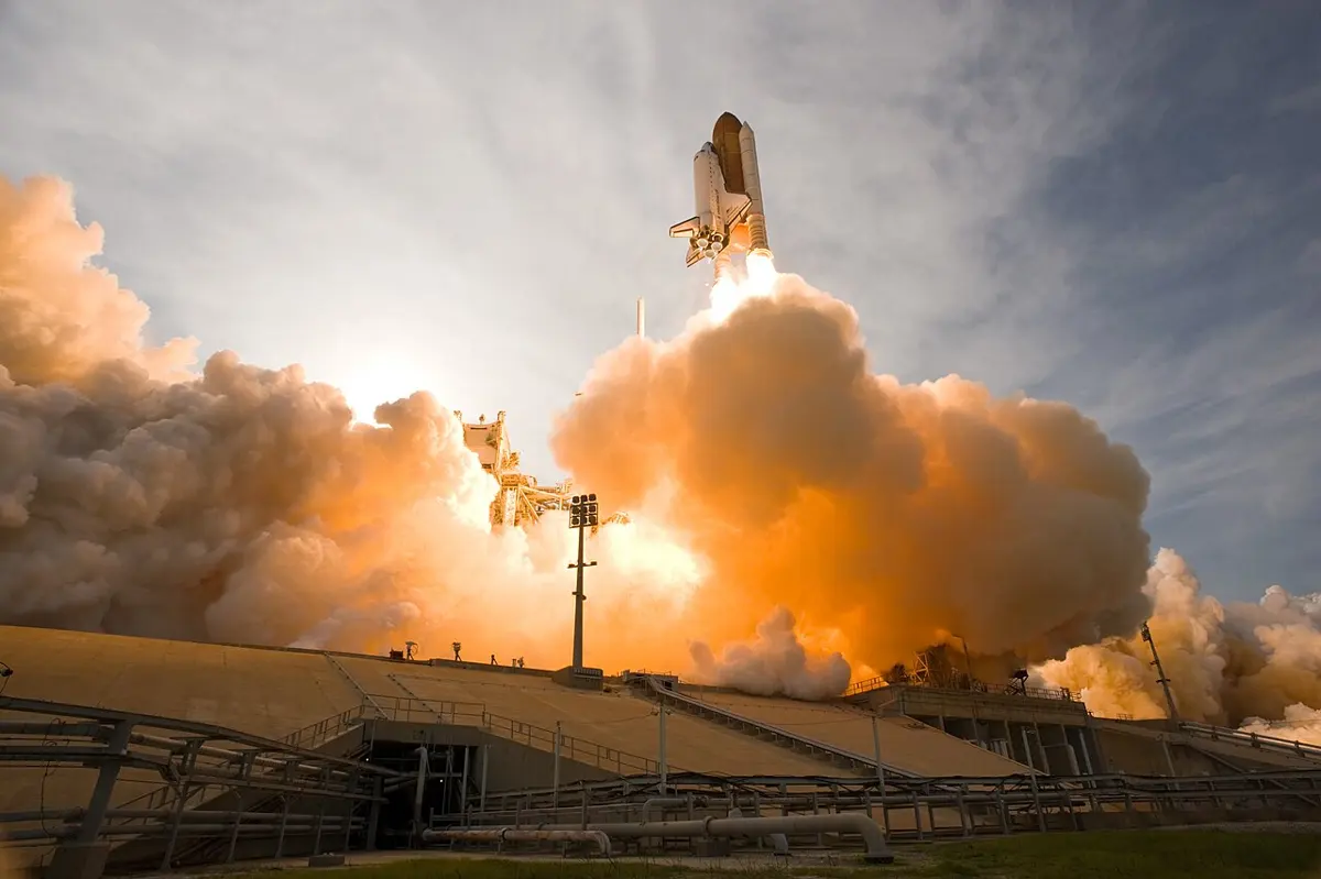 A space shuttle with its large tank and two white boosters rising on a bright flame above huge clouds of orange-lit smoke at the launch pad