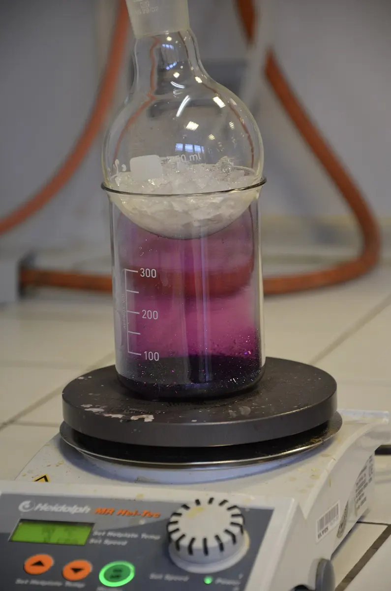 A glass beaker on a laboratory hot plate, filled with violet vapor, with a round flask full of ice sitting in its mouth