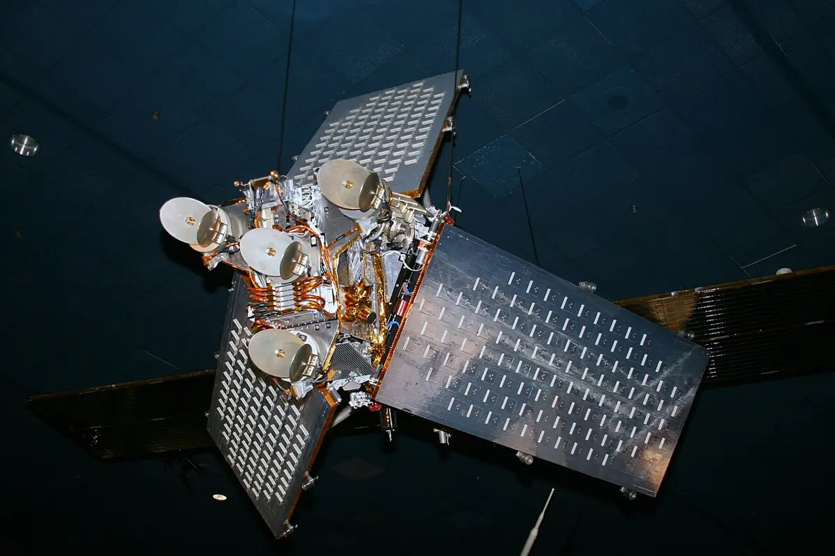 A satellite hanging under a dark museum ceiling: a box-shaped body with four round dish antennas and gold-colored parts, large flat silver panels with rows of small slots, and dark solar panels behind