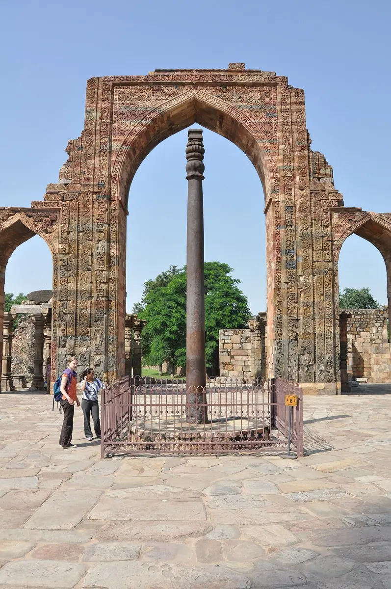 A tall, dark, slender metal pillar inside a low railing, standing in a stone courtyard in front of a ruined arch of carved red stone, with two visitors beside it