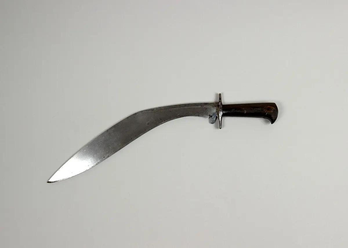 A curved knife with a broad steel blade bending forward and a dark handle with a metal guard, on a plain gray background