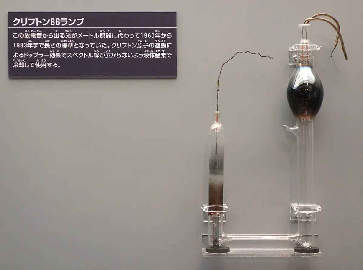 A glass apparatus on a museum wall: a thin upright tube on the left joined at the bottom to a taller tube with a dark round bulb on the right, wires coming out of the tops, beside a sign in Japanese