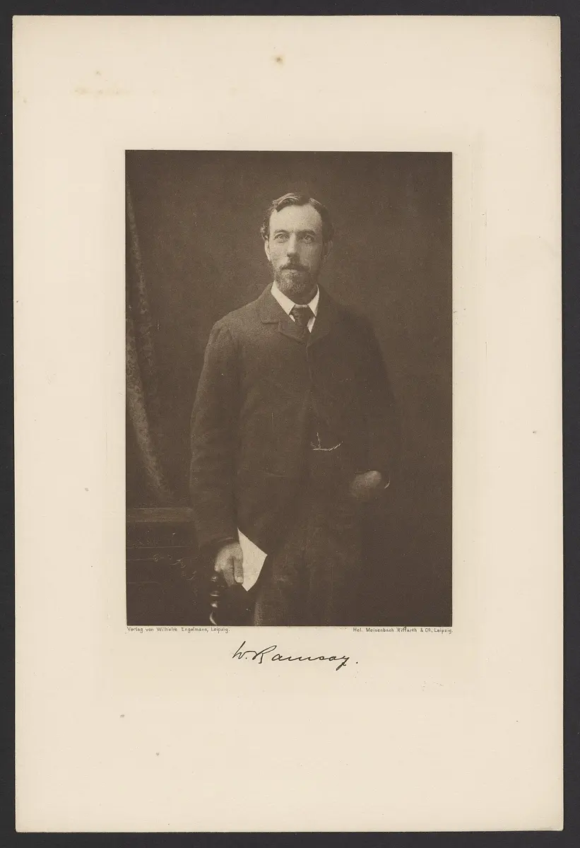 An old brown-toned photograph of a man with a short beard and mustache, standing in a dark suit and holding some papers, with his signature, W. Ramsay, below