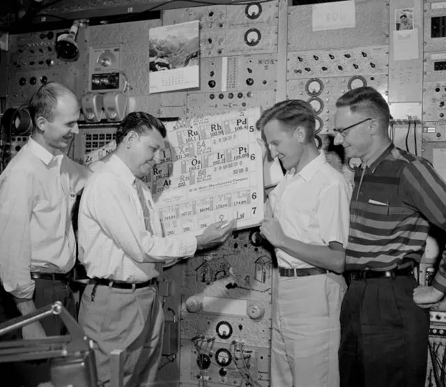 A black-and-white photo of four smiling men in shirts in front of a wall of dials and switches; one writes with a pen on a large printed periodic table while the others watch