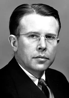 A black-and-white portrait of a man with combed-back hair and rimless glasses, in a dark suit and tie