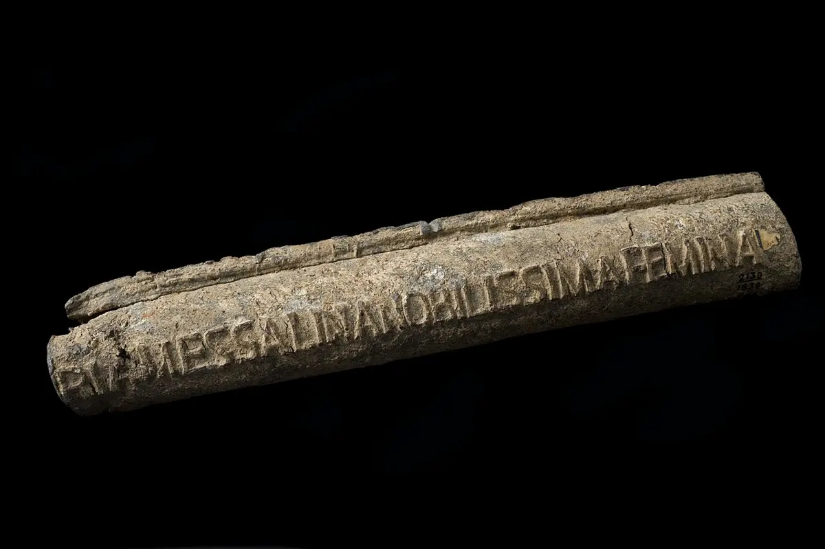 A short length of old gray metal pipe on a black background, with a seam along its top and raised Latin capital letters along its side