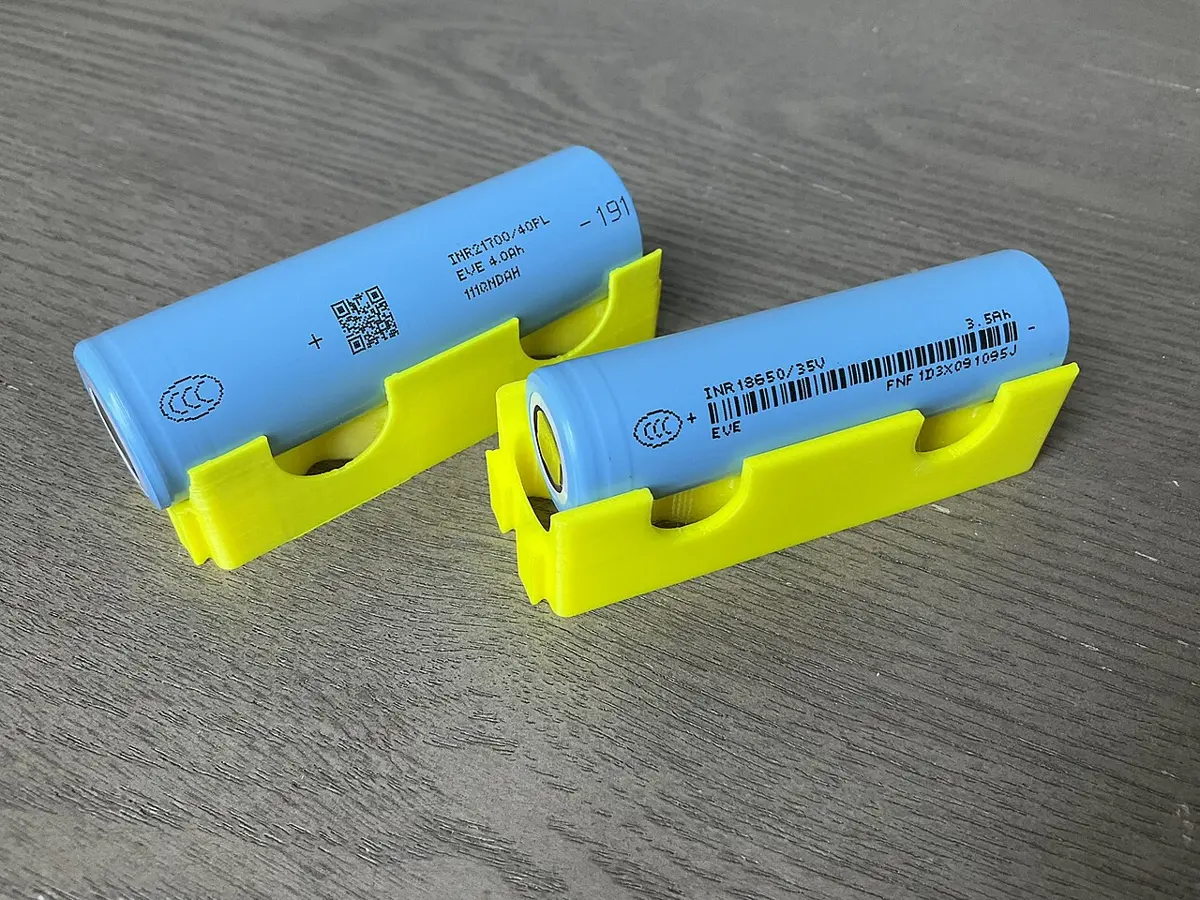 Two light-blue cylindrical battery cells resting in yellow holders on a wooden table, one a little fatter than the other
