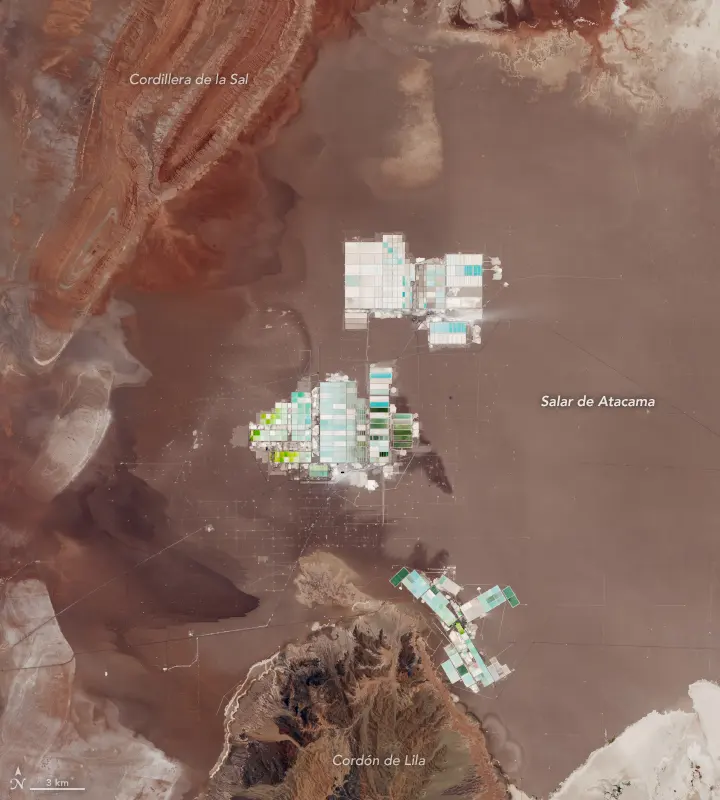 A satellite view of a brown salt flat beside red-brown hills, with three patches of rectangular ponds in white, turquoise and green