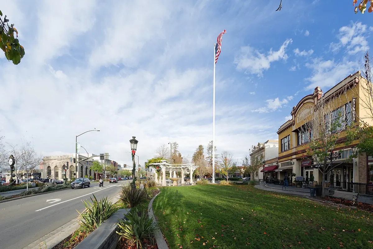 A wide street in a small town on a sunny day, with a tall flagpole on a grassy strip in the middle, old two-storey shop buildings and a street clock