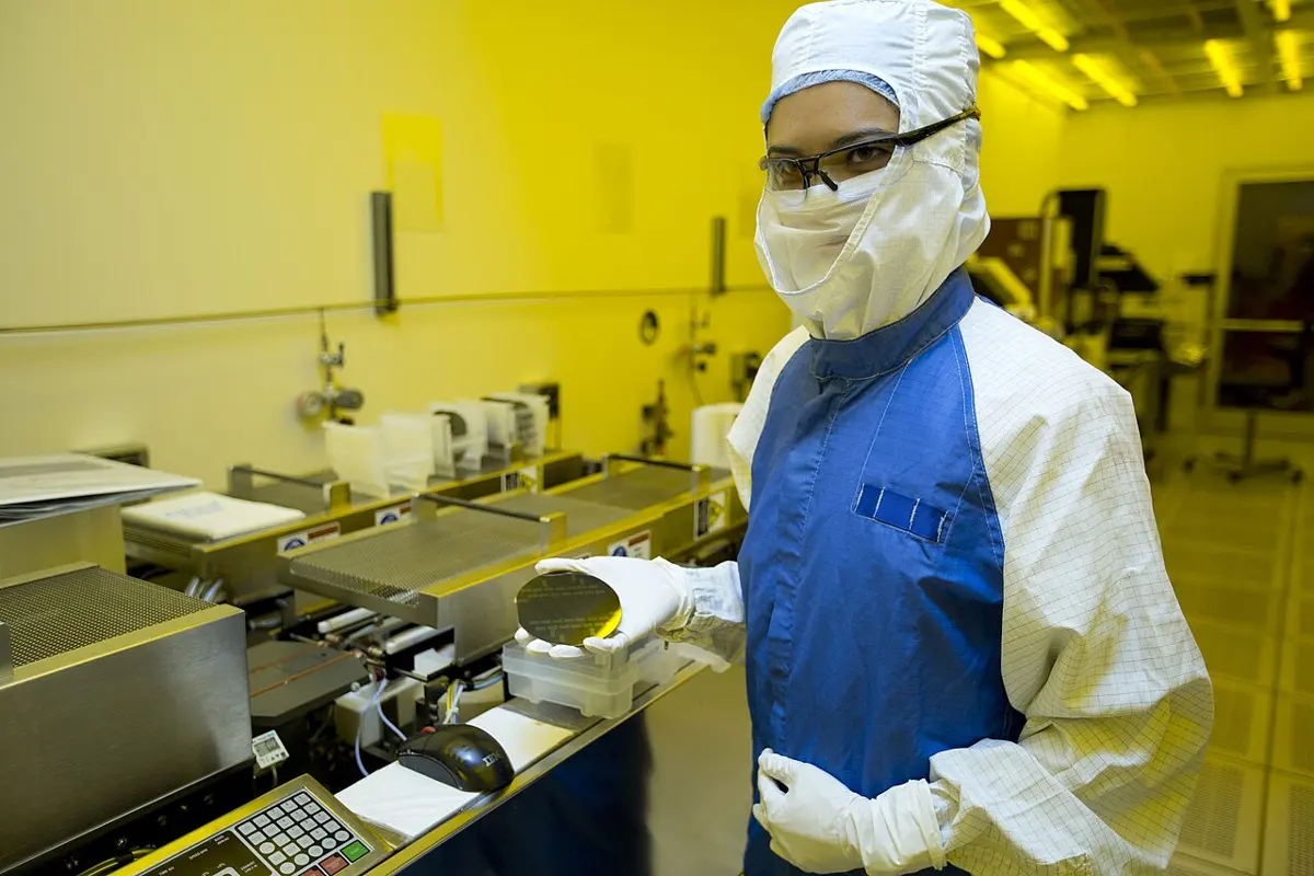 A scientist in a white cleanroom suit, hood, face mask and gloves holds up a round silicon wafer in a room lit with yellow light