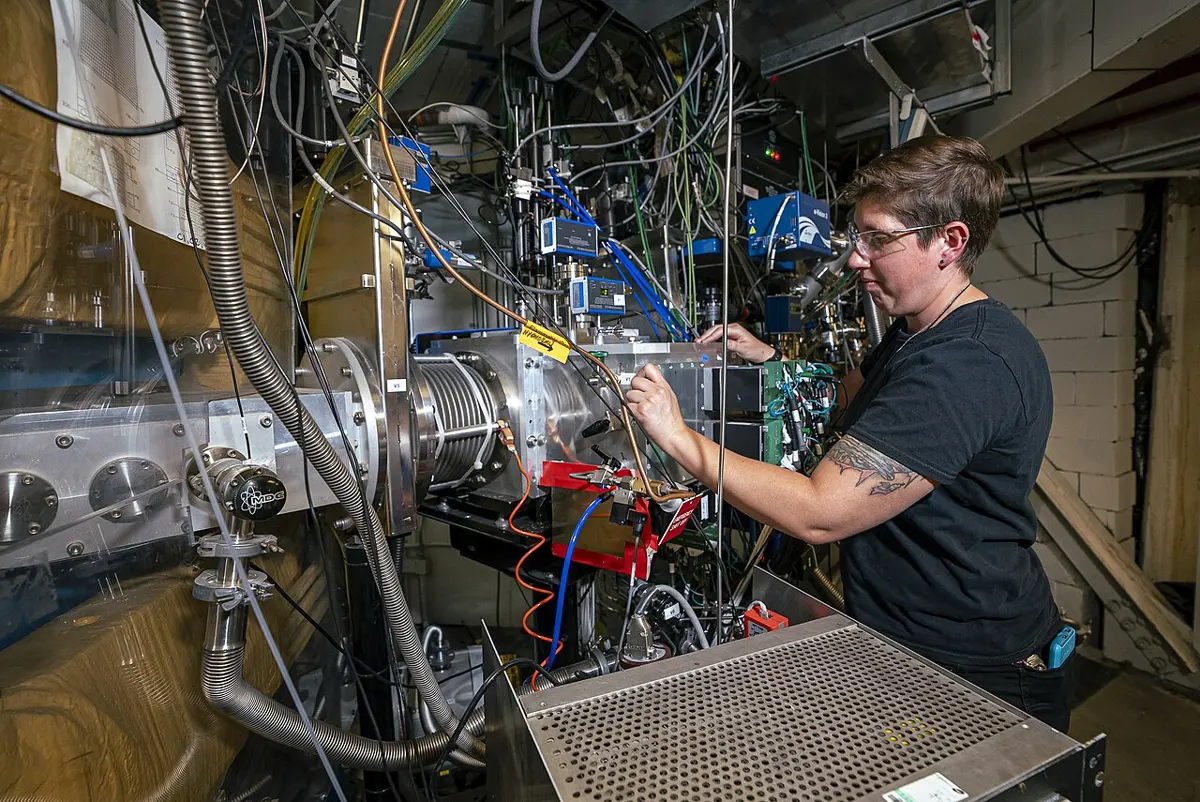 A scientist in a black T-shirt and safety glasses adjusts a steel machine crowded with vacuum pipes, valves and cables, in a basement room
