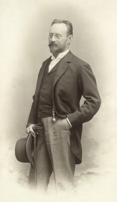 An old sepia photograph of a standing man with glasses and a beard, in a dark jacket and striped trousers, holding a hat and a walking stick