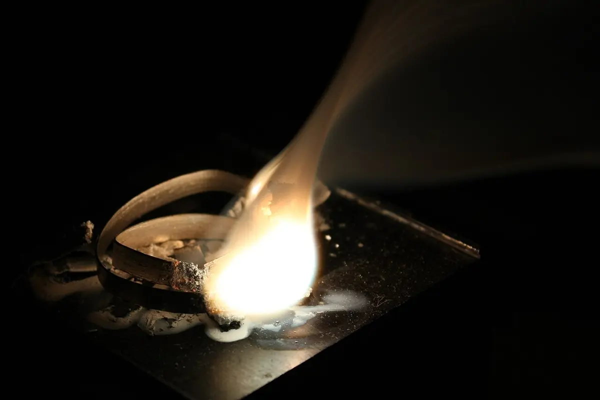 A coil of metal ribbon on a dark plate, burning at one end with a blinding white flame and a plume of white smoke, with white ash beside it