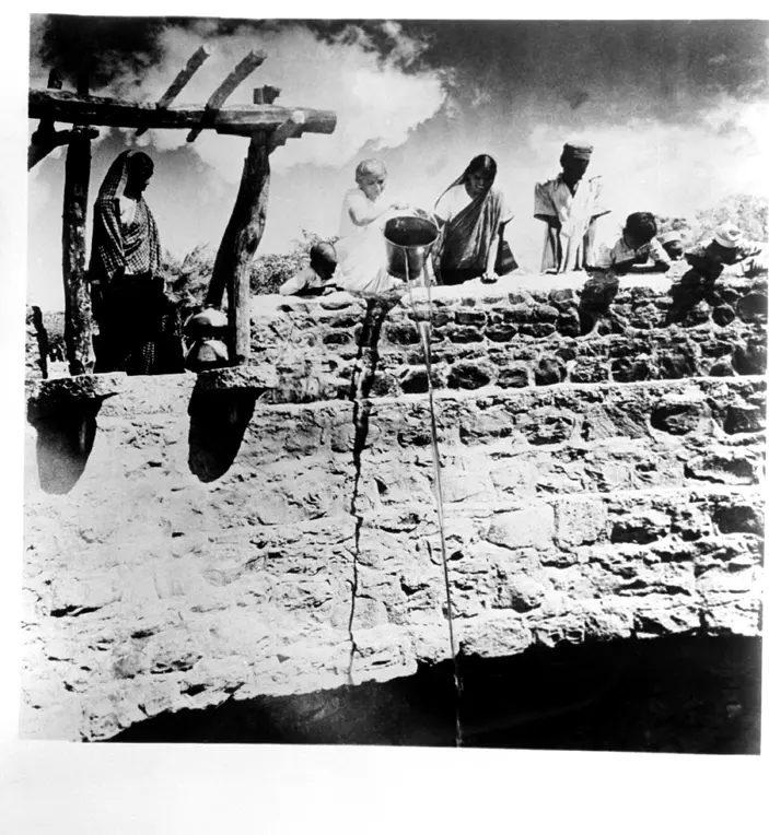 An old black-and-white photo of a stone village well: a woman on its wall tips a bucket, and a thin stream of liquid falls into the well while women and children watch