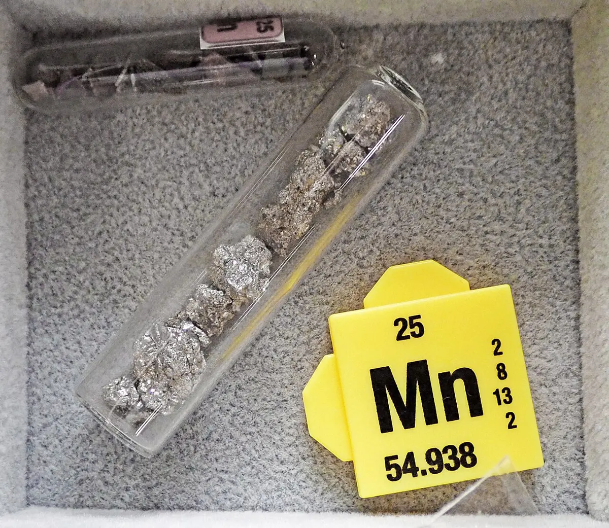 Crumpled, shiny, silvery-gray pieces of metal in a glass tube, beside a yellow card that reads Mn, 25 and 54.938