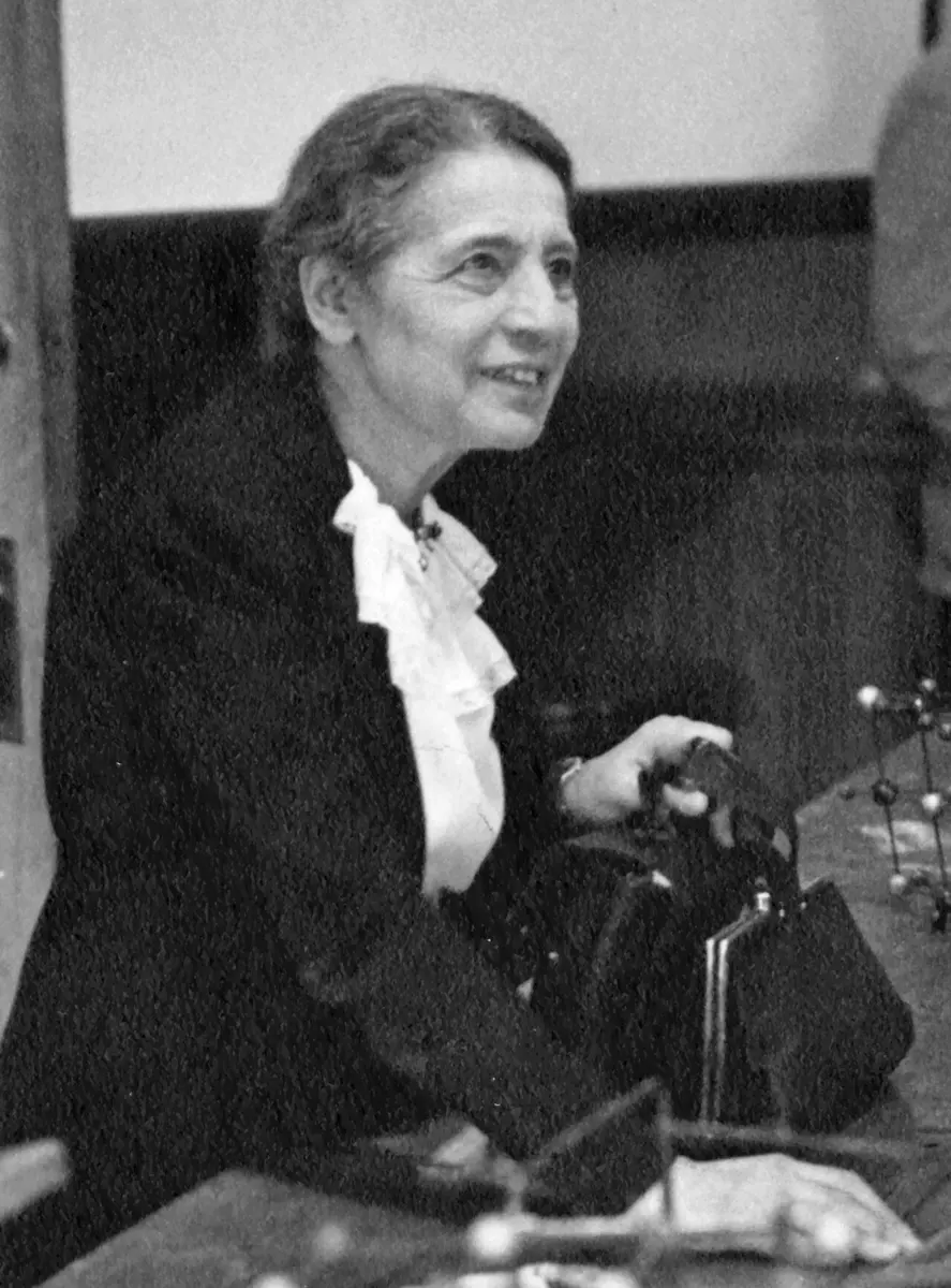 A black-and-white photo of an older woman with gray hair, in a dark jacket and a white frilled blouse, smiling, one hand on a bag on a lecture bench, with a small ball-and-stick model beside her