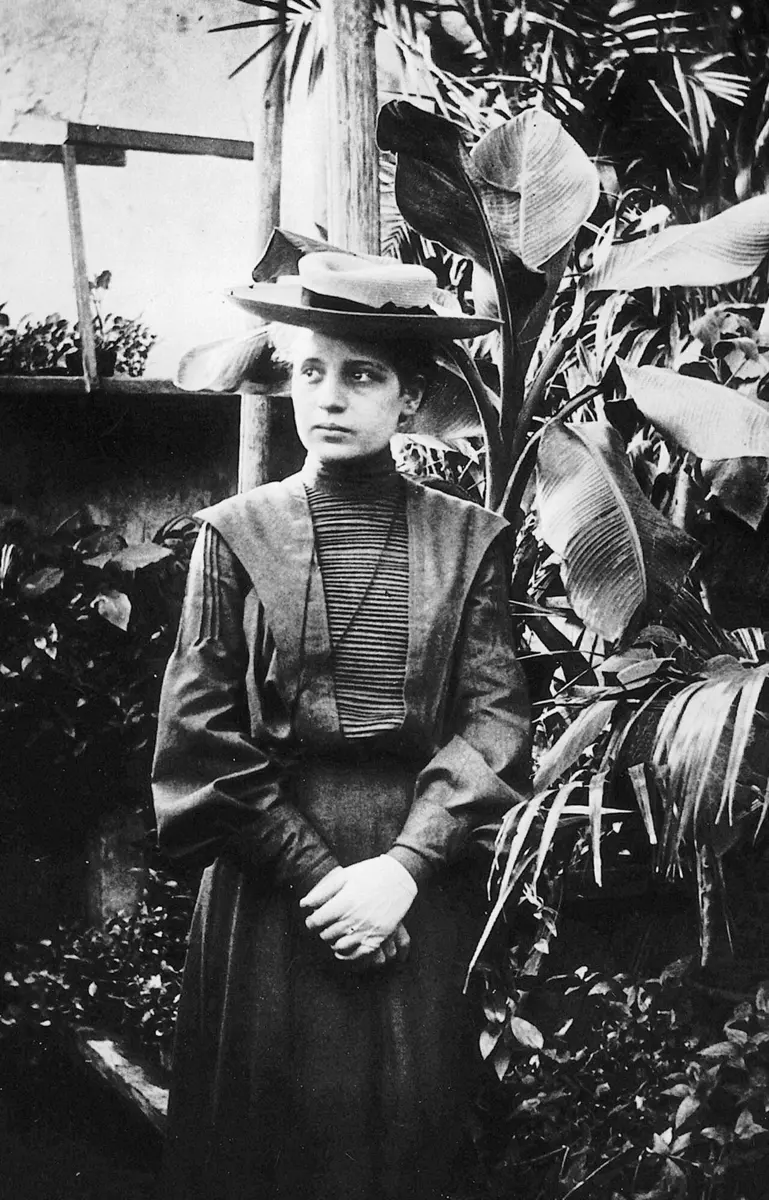 An old black-and-white photo of a young woman in a long dark dress and a straw hat, standing among large potted plants