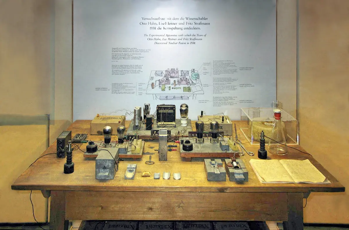 A wooden table in a museum display case, covered with small instruments, wires, glass vessels and paper notes, under a wall panel of text