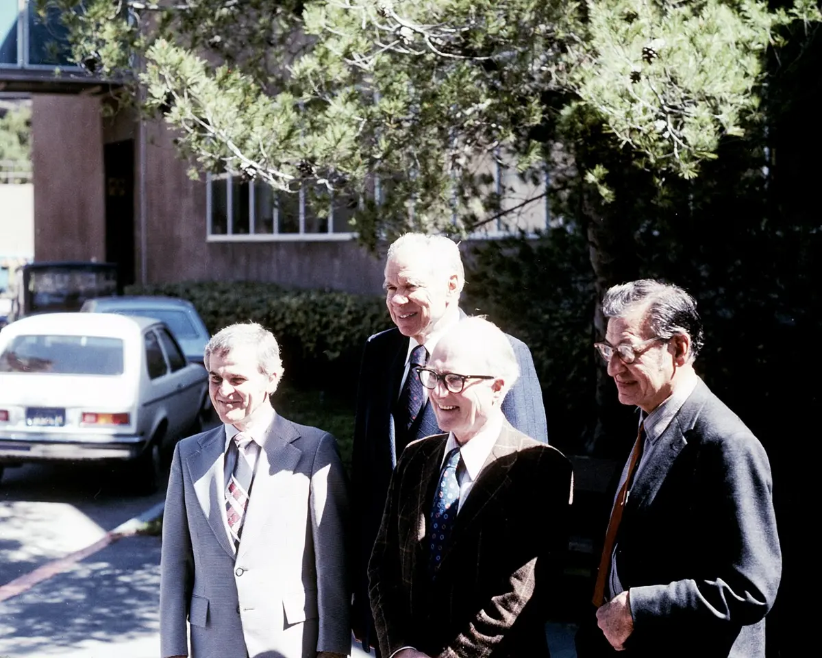 Four smiling older men in suits standing outdoors in the sun, under a pine tree, with a building and parked cars behind them